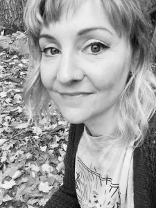 Black and white photo of Beth Starling, showing a young white woman with fair hair and a nose ring, wearing a t shirt and cardigan. There are leaves in the background. 